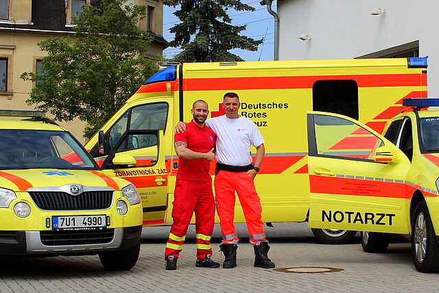 Ein Tschechischer und ein Deutscher Mitarbeiter des Rettungsdienstes stehen Arm in Arm vor ihren Einsatzwagen.