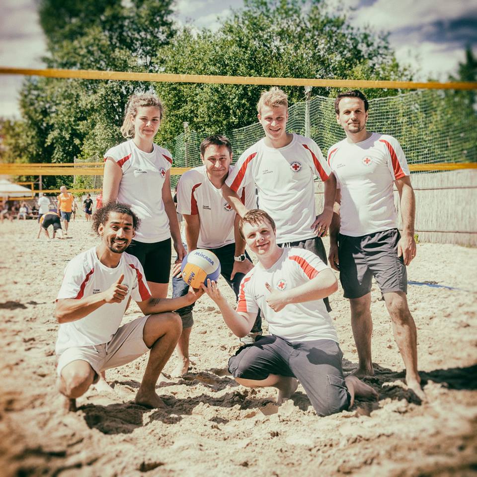 Sechs Volleyballspielerinnen und Volleyballspieler stehen auf einem Strandvolleyballfeld und lächeln in die Kamera. Zwei der Spieler halten den Volleyball in den Händen.
