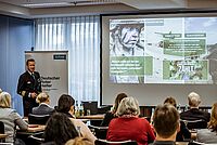 Ein Mitarbeiter der Bundeswehr steht neben der leinwand mit seiner Präsentation vor den Teilnehmern und berichtet von seinem Thema.