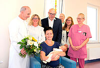 Claire Krumbiegel brachte die 1000. Geburt des DRK Krankenhauses hinter sich und einen kleinen Jungen auf die Welt. Auf dem Bild posieren Ärzte, Schwestern, die Mutter mit ihrem Baby sowie das DRK Vorstandsmitglied Kerstin Trautmann. Insgesamt sechs Personen stehen um die Mutter, die mit ihrem Sohn in der Mitte sitzt.