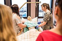 Die Dozentin zeigt den Auszubildenden am Beispiel einer Puppe pflegerische Tätigkeiten einer Krankenpflegehilfe.