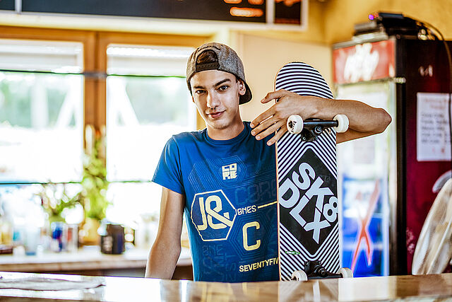 Ein Jugendlicher steht mit einem Skateboard in der Hand hinter einer Bar eines Jugendzentrums.