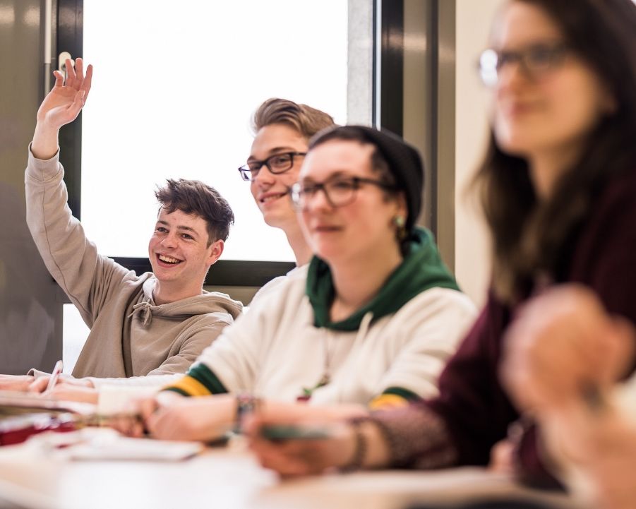 Mehrere Schülerinnen und Schüler sitzen in einem Schulungsraum. Ein Schüler meldet sich durch ein Handzeichen, um etwas zu sagen.