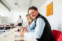Mehrere Personen sitzen in einem Konferenzraum. Ein Mann dreht sich zur Kamera und lächelt. Im Hintergrund sieht man einen weiteren Mann, der zu den Personen spricht.